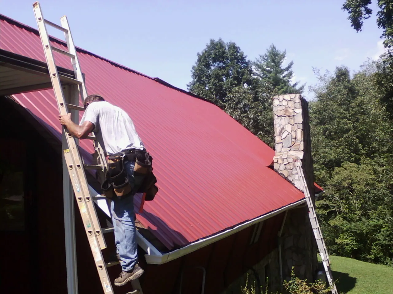 Roofer on a ladder beside a red metal roof and stone chimney — Soffit & Fascia Repair by Roofing Brothers in Upperco